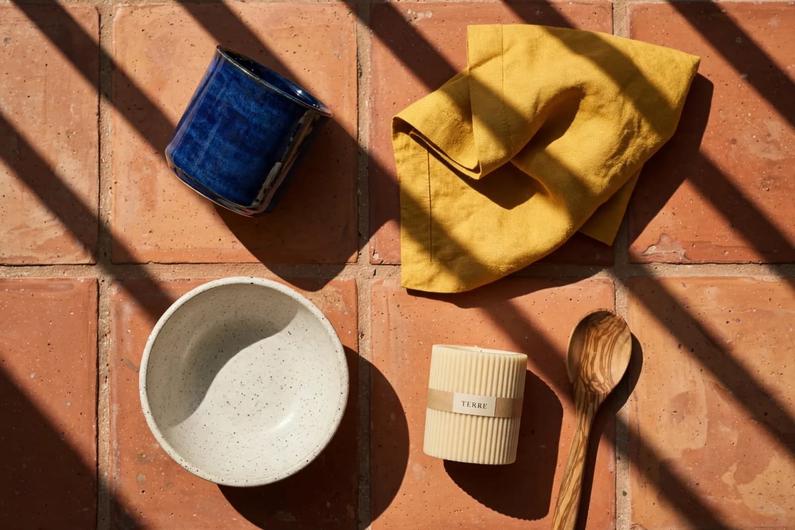 A homewares range on sunlit terracotta tiles: a cobalt cup, a mustard napkin, a bowl, a Terre candle and a wooden spoon